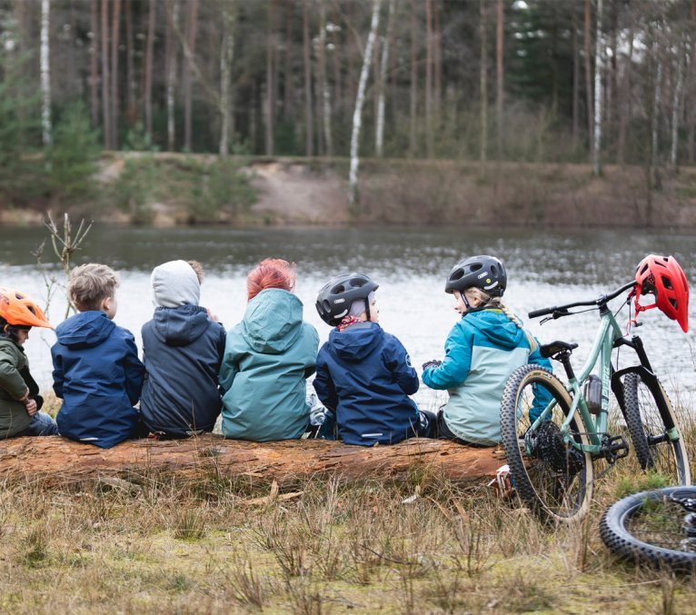 Kinder sitzen auf einem Baum an einem See. Sie sind mit ihren Mountainbikes unterwegs. 