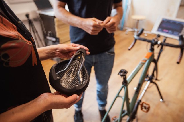 Eine Person hält einen schwarzen, thermoformbaren Fahrradsattel von Reform in den Händen, während eine andere Person im Hintergrund steht. Im Hintergrund ist ein Fahrrad auf einem Ständer sowie ein Laptop auf einem Tisch zu sehen.