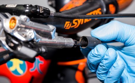 Gros plan sur une durite de frein de vélo tandis qu’une personne portant des gants bleus utilise un outil pour desserrer ou installer le raccord.