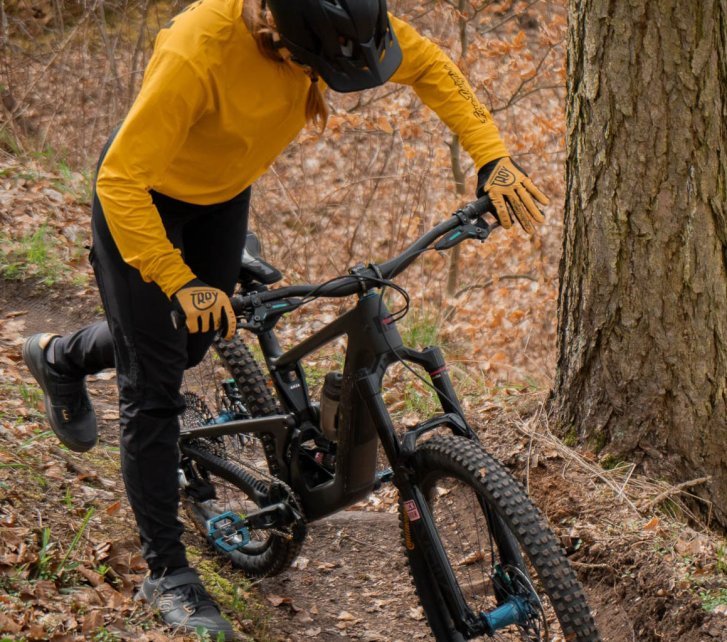 Una mujer con maillot ocre y casco oscuro de MTB se sube a una bicicleta de montaña oscura.