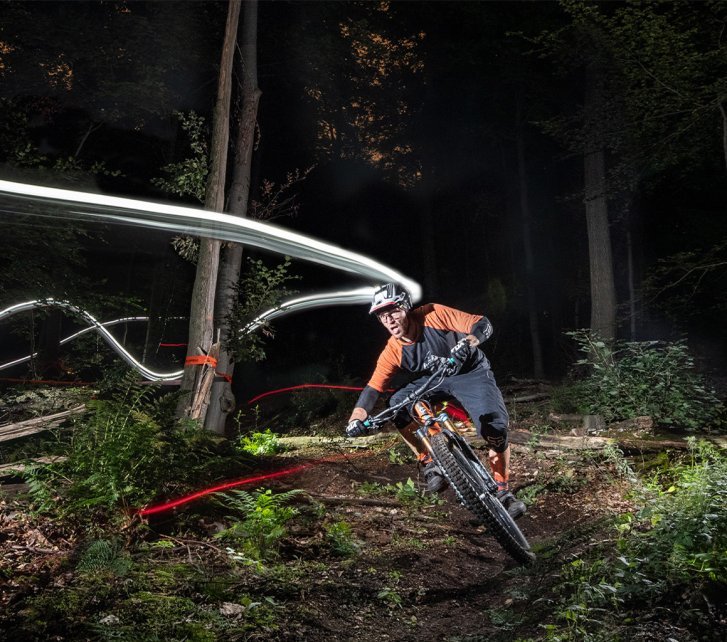 Un ciclista de montaña pedalea de noche en un sendero del bosque, iluminado por una luz en el casco, con trazos de luz marcando el camino y árboles rodeando el entorno.