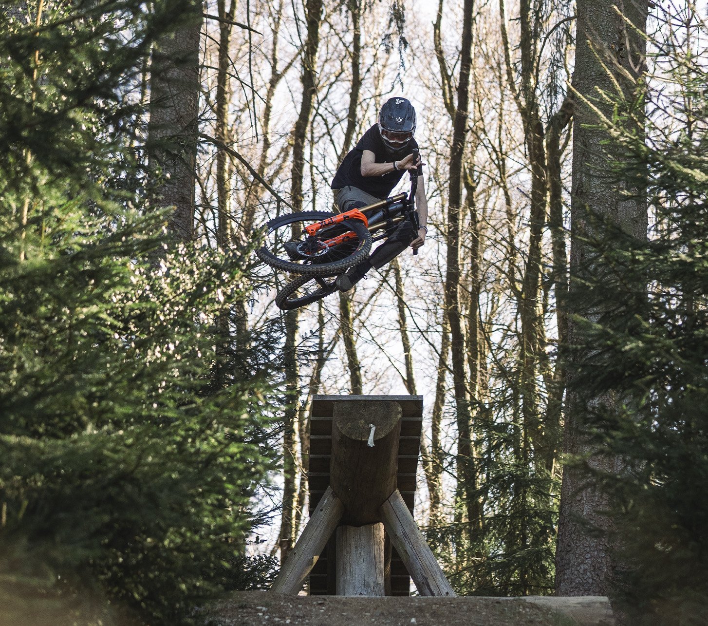 Mountainbiker mit Helm und Schutzkleidung springt über einen großen Holzkicker in einem Wald; das Bike ist in der Luft quer gedreht.