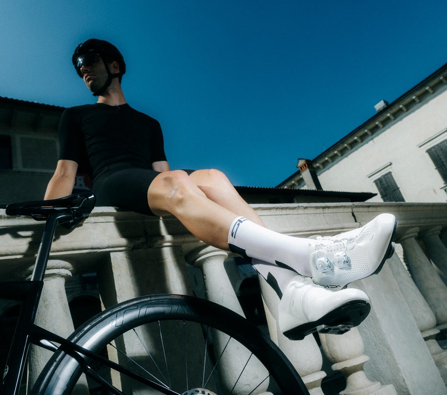 Rennradfahrer sitzt mit Rennrad auf einer Balustrade und trägt weiße SIDI Rennradschuhe.