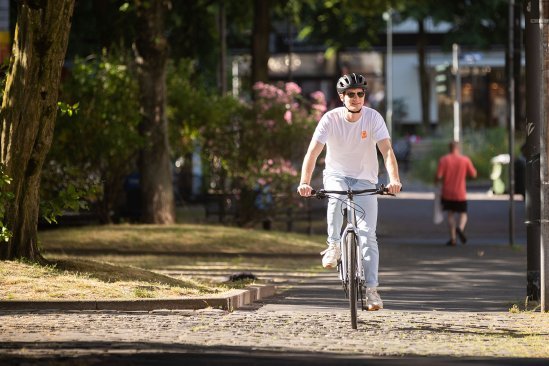 A person rides a bicycle on a paved bike path in an urban setting, surrounded by trees and a relaxed atmosphere, while another person goes in the background.