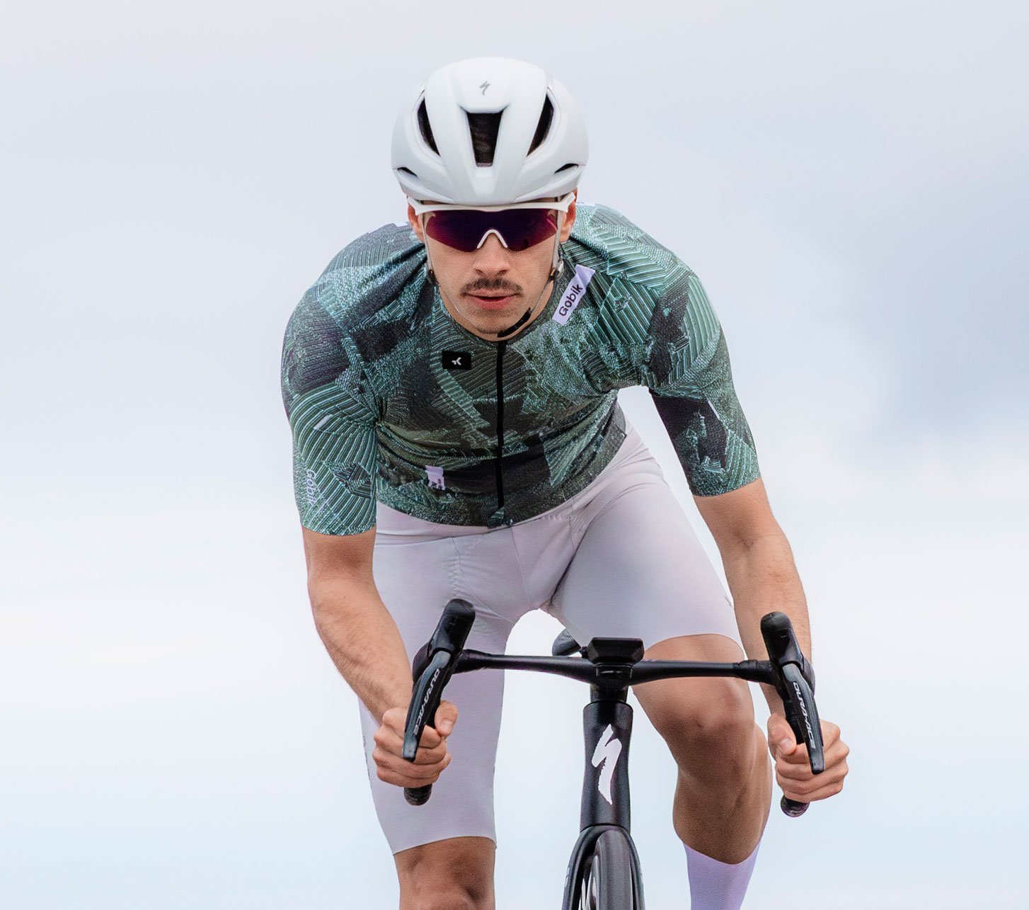 Ciclista de carretera en posición aerodinámica con casco blanco y gafas deportivas, vistiendo maillot verde Gobik y culotte blanco sobre bicicleta de carretera moderna – ropa de ciclismo de alto rendimiento