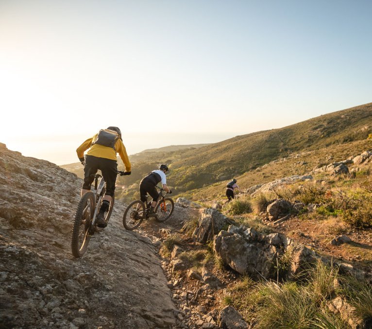Drei E-Mountainbiker befahren einen felsigen Trail im Gegenlicht. Wir sehen die drei von hinten, die Abendsonne taucht alles in ein goldenes Licht.