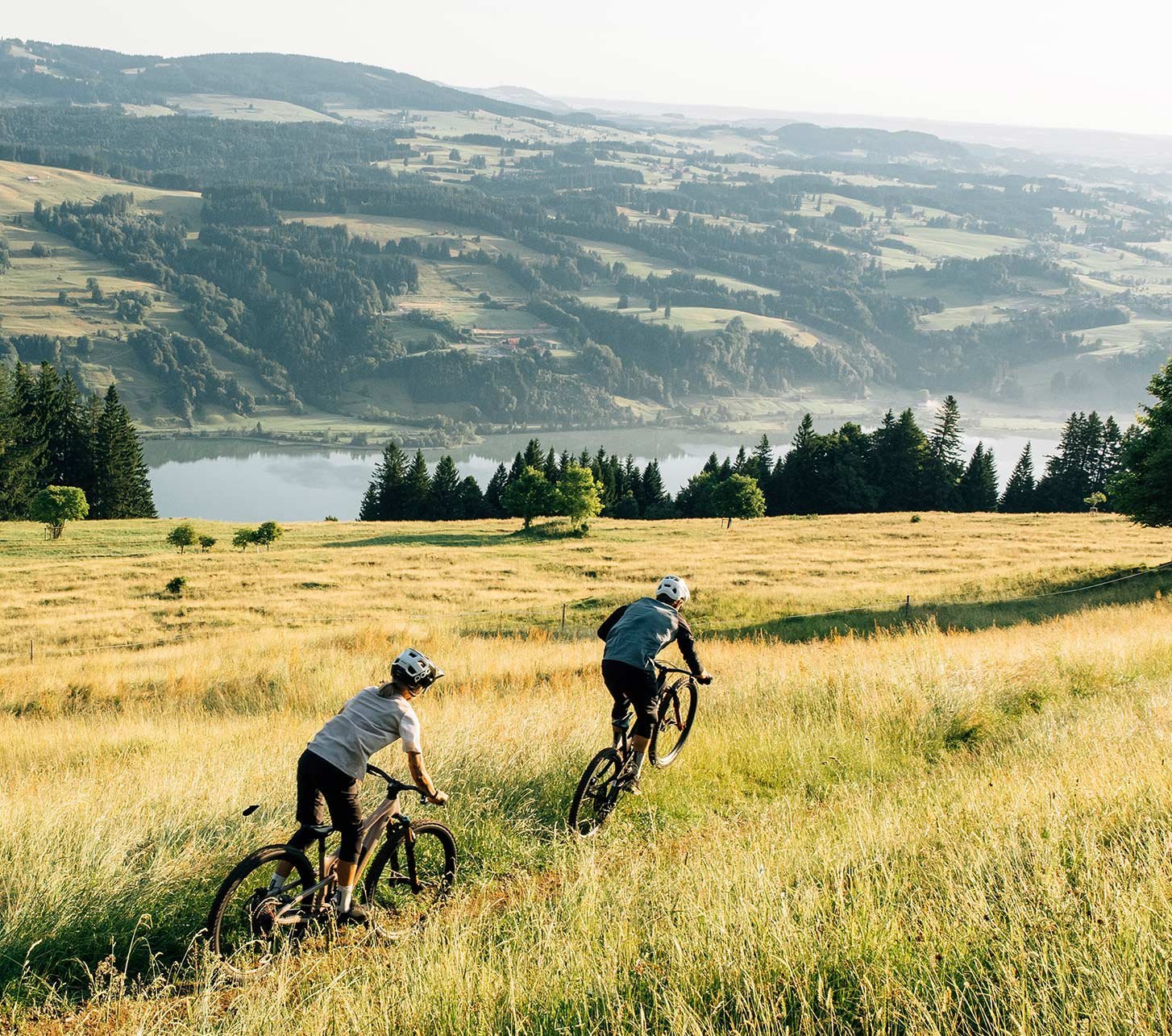 Zwei Mountainbiker in VAUDE Fahrrabekleidung fahren mit ihren Mountainbikes über eine hohe Feldwiese - im Hintergrund Alpines Talgelände und ein Bergsee.