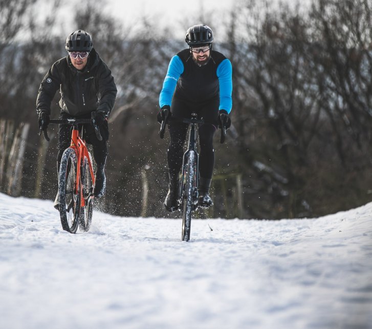 Dos hombres en gravel bikes rodando hacia la cámara en un paisaje nevado.