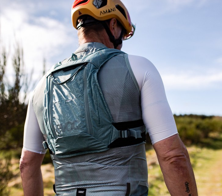 Ein Mann in Fahrradkleidung von schräg hinten, wir sehen nur den Oberkörper vor dem unscharfem Hintergrund einer Naturlandschaft mit grüner Vegetation und blauem Himmel. Der Mann trägt einen blaugrauen enganliegenden Trinkrucksack 