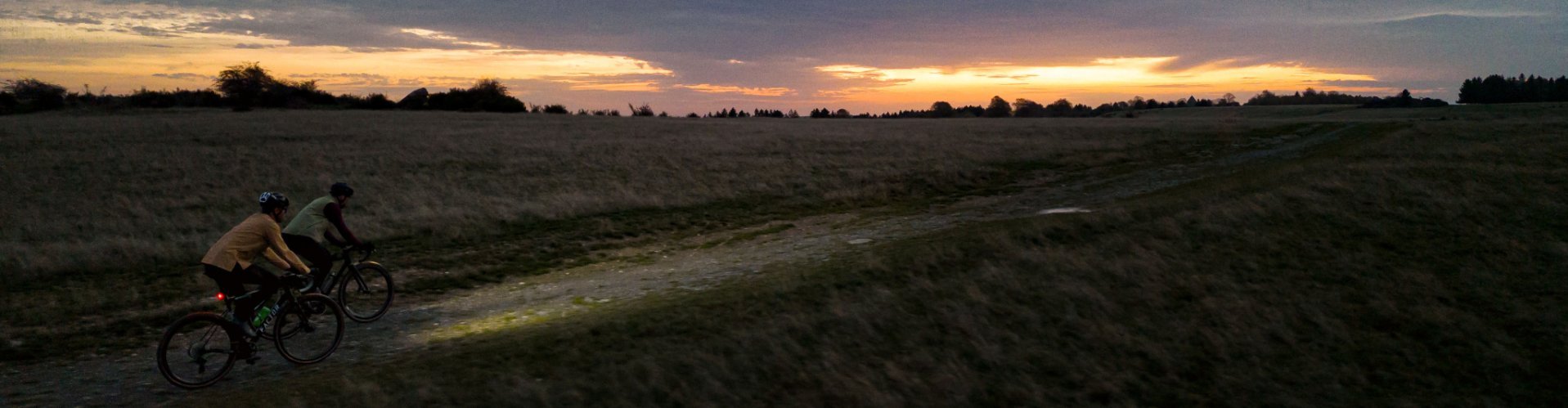 Dos ciclistas con las luces encendidas recorren un camino rural al atardecer.
