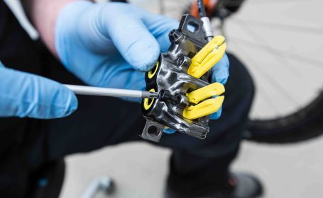 Close-Up-Foto eines Magura Bremssattels, gehalten von einer Hand in blauen Einweghandschuh. Gelbe Distanzblöcke sind eingesetzt, die Bremsbelag-Halteschraube wird wieder eingesetzt.