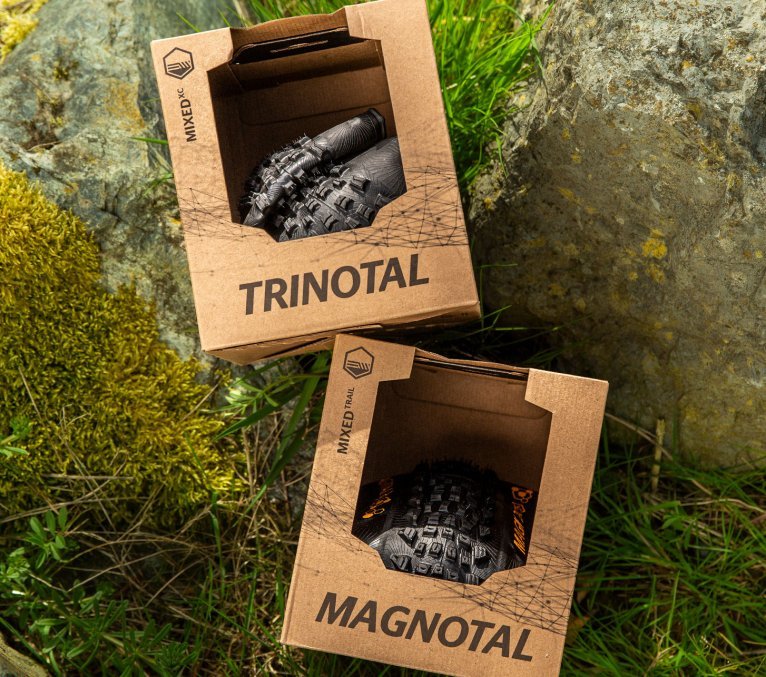 Zwei Continental Fahrradreifen in Kartonverpackungen mit den Namen Trinotal und Magnotal liegen zwischen Steinen und Gras im Outdoor-Setting.