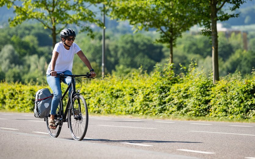 A woman rides a bicycle with a pannier bag on a road, surrounded by a green, rural environment with trees and hills in the background.