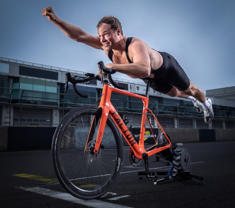 A road bike with an indoor trainer is set up on the starting line of a racetrack. A half-naked man lies belly-down on the saddle in a Superman pose.