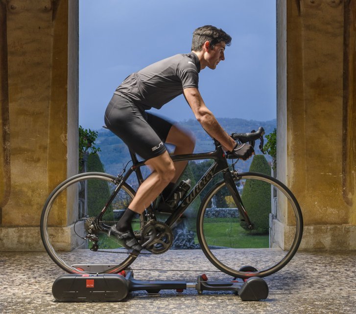 A man trains on a road bike set up on free rollers in front of a Mediterranean backdrop.