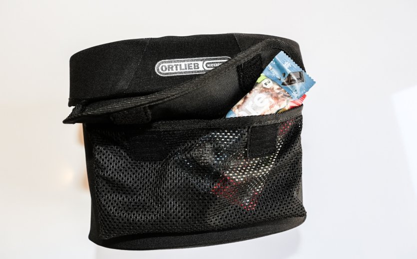 A black Ortlieb bicycle bag with a black mesh pocket on the side, containing a visible energy gel bar. The bag is labeled "Ortlieb Waterproof."