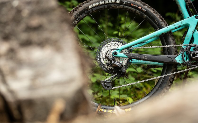 Detrás de una piedra difuminada se ve el lado de la transmisión de una bici de montaña de color turquesa.