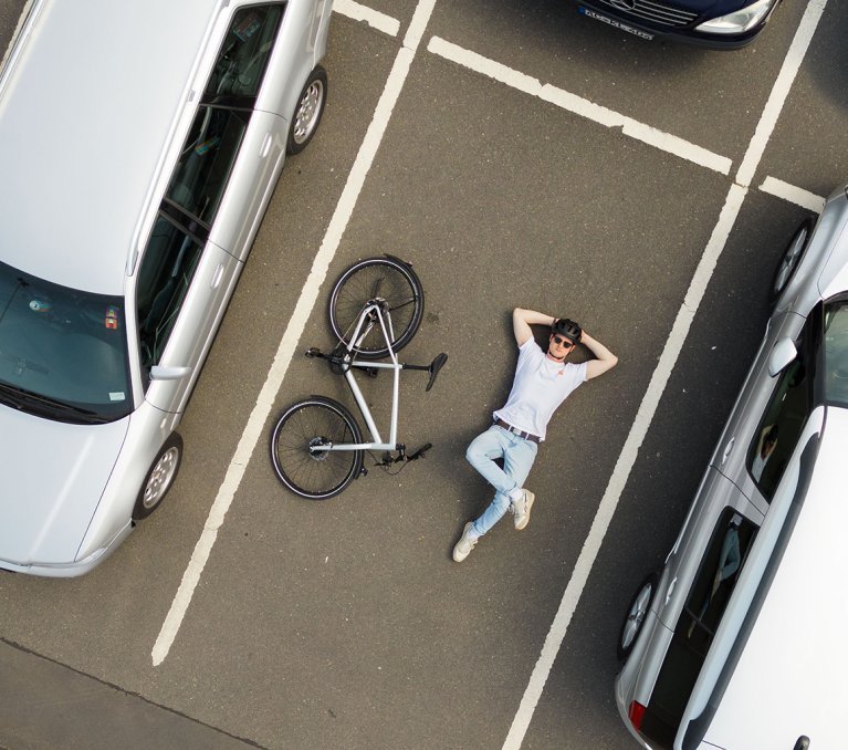 A person is lying relaxed on the ground of a parking lot next to a bicycle, which is also on the ground, between two parked cars.