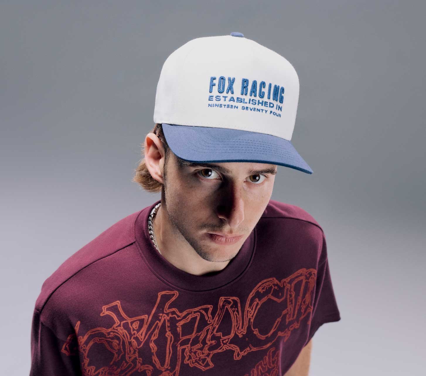 Homme portant une casquette Fox Racing blanche et bleue et un t-shirt Fox Racing bordeaux sur fond gris.