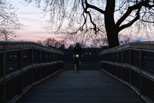 Eine Person auf einem Fahrrad fährt über eine hölzerne Brücke auf die Kamera zu. Es ist fast Nacht und man erkennt fast nur die neongelben Hand- und Überschuhe sowie das Licht der Frontleuchte am Fahrrad.