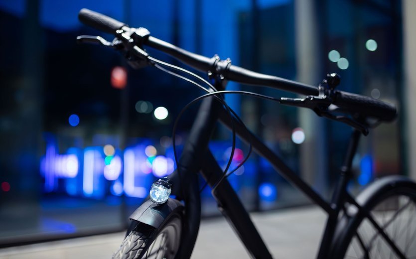 Gros plan d’un vélo urbain au crépuscule avec un phare avant alimenté par dynamo allumé ; guidon et roue avant visibles, lumières de la ville floues en arrière-plan.