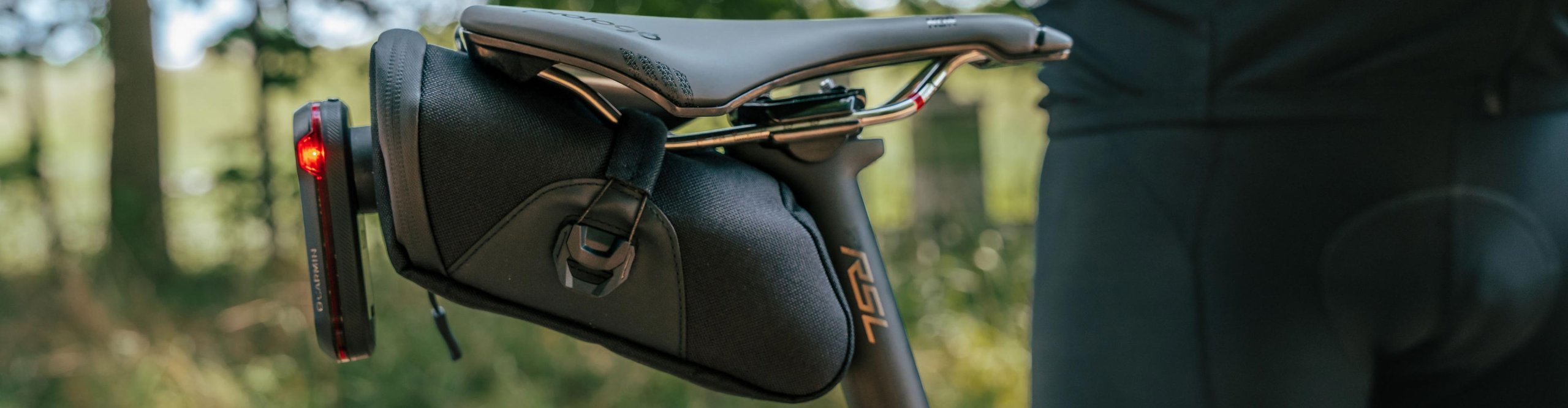 KOM Cycling saddle bag mounted beneath a road or gravel bike saddle with rear light, photographed outdoors in the forest