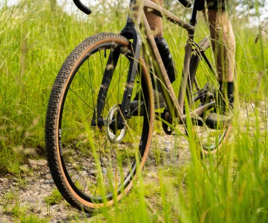 Roue avant d’un vélo gravel sur chemin caillouteux, pneu à crampons marqués et flanc beige en action dans un environnement verdoyant.