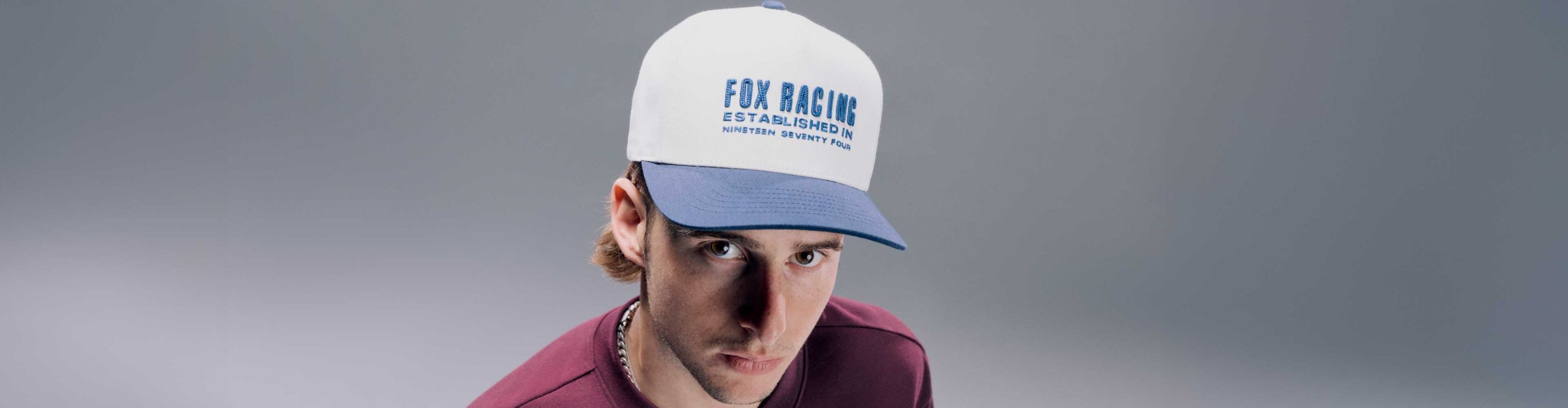 Homme portant une casquette Fox Racing blanche et bleue et un t-shirt Fox Racing bordeaux sur fond gris.