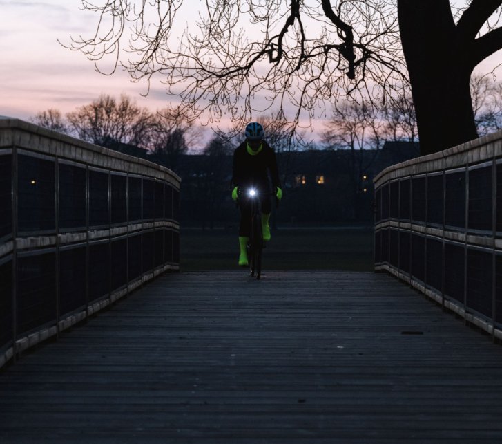 Eine Person auf einem Fahrrad fährt über eine hölzerne Brücke auf die Kamera zu. Es ist fast Nacht und man erkennt fast nur die neongelben Hand- und Überschuhe sowie das Licht der Frontleuchte am Fahrrad.
