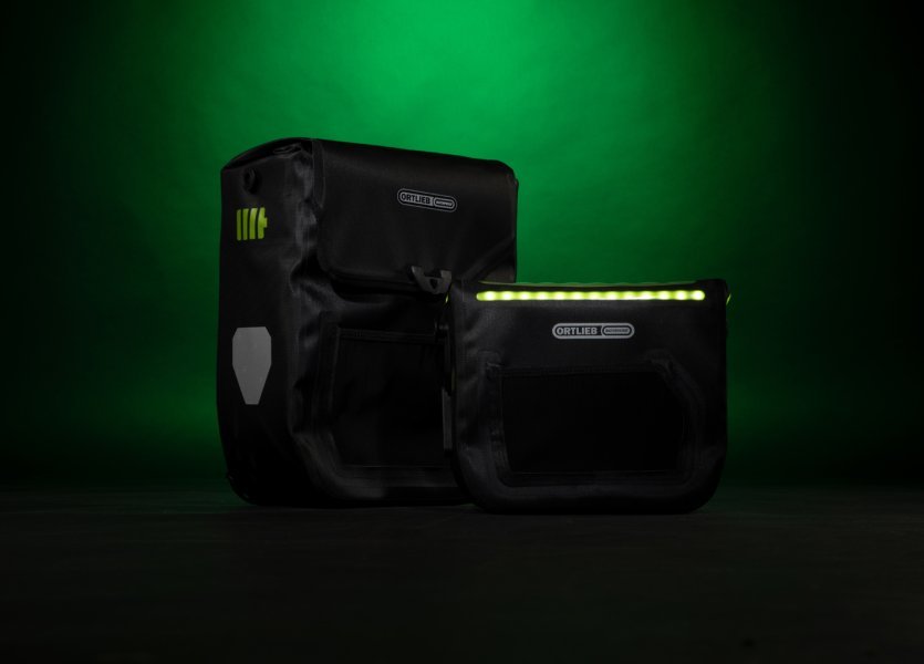 Zwei ORTLIEB Fahrradtaschen – die beleuchtete E-Glow Lenkertasche und die E-Mate Gepäckträgertasche – vor grünem Hintergrund