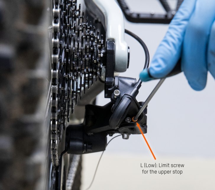 The rear derailleur can be seen from behind. A hex wrench is put on the outer stop. An orange arrow marks the L-screw. 