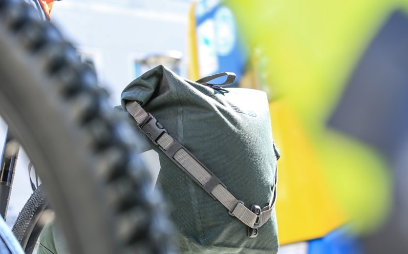 Nahaufnahme einer grünen Ortlieb Back-Roller Urban Fahrradtasche, die an einem Fahrrad befestigt ist. Im Vordergrund ist ein Fahrradreifen sichtbar, im Hintergrund ein verschwommener Blick auf eine bunte Umgebung.