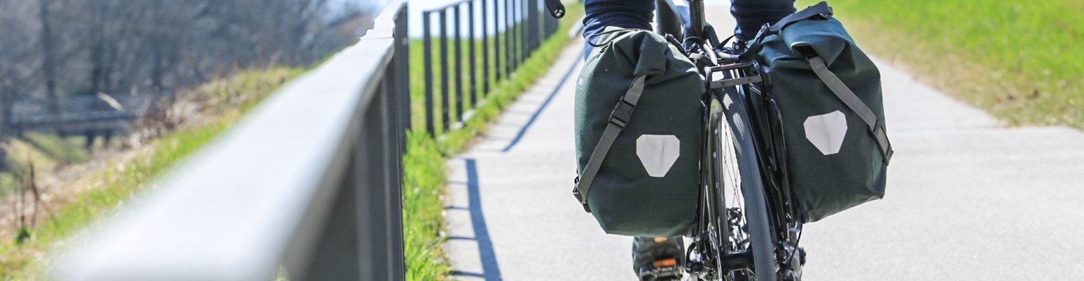 Ein Fahrradfahrer in einer roten Jacke fährt auf einem Radweg, mit zwei grünen Ortlieb Back-Roller Urban Fahrradtaschen, die an beiden Seiten des Hinterrads befestigt sind. Im Hintergrund sind eine grüne Wiese und kahle Bäume zu sehen.