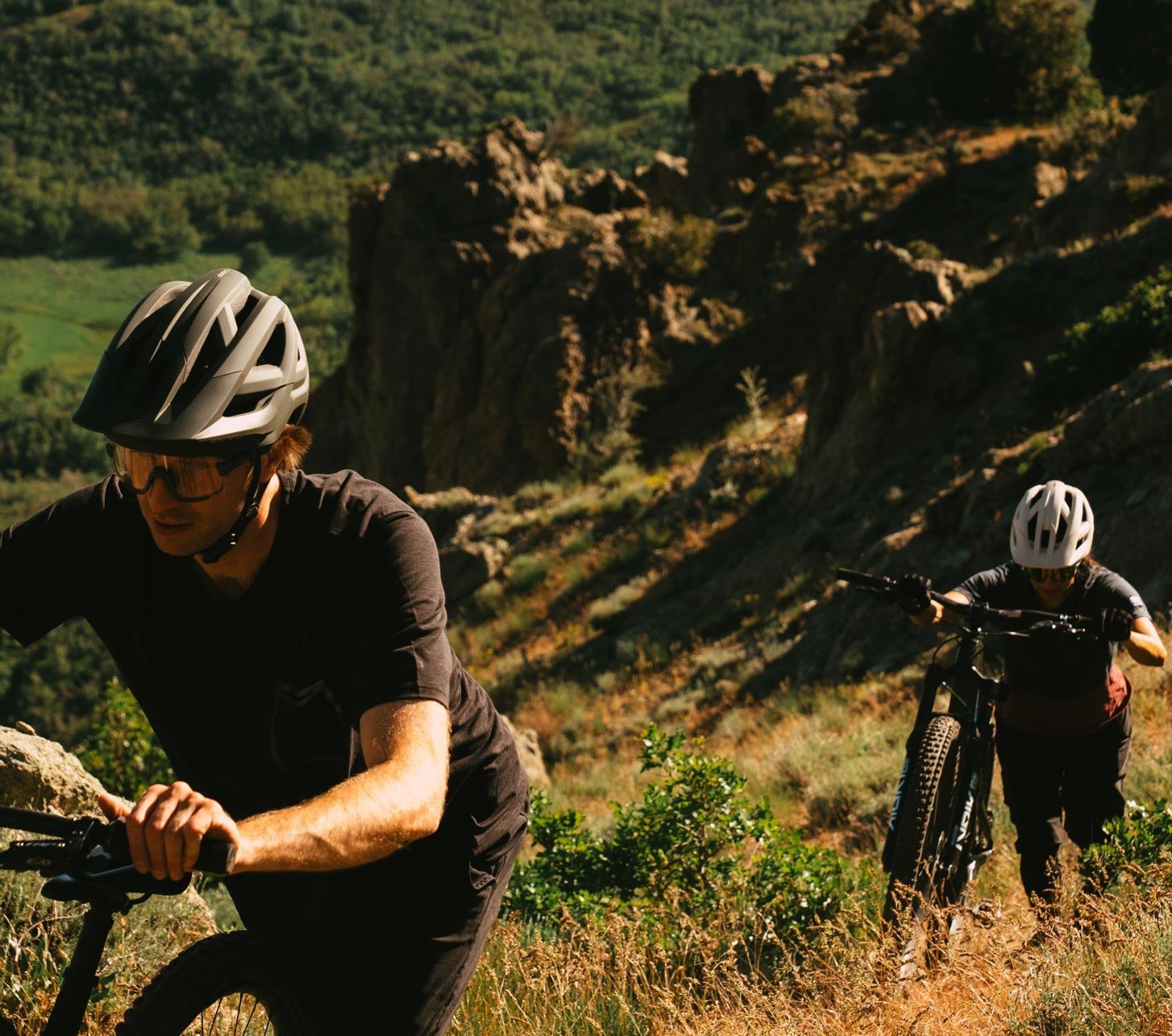 Zwei Mountainbiker mit Giro Mountainbike Helmen schieben ihre Bikes auf einem steilen, felsigen Trail über einem grünen Tal.