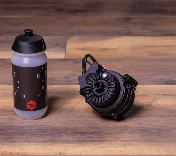 A TQ HPR50 e-bike drive lies on a dark wooden floor. A bike water bottle stands next to it