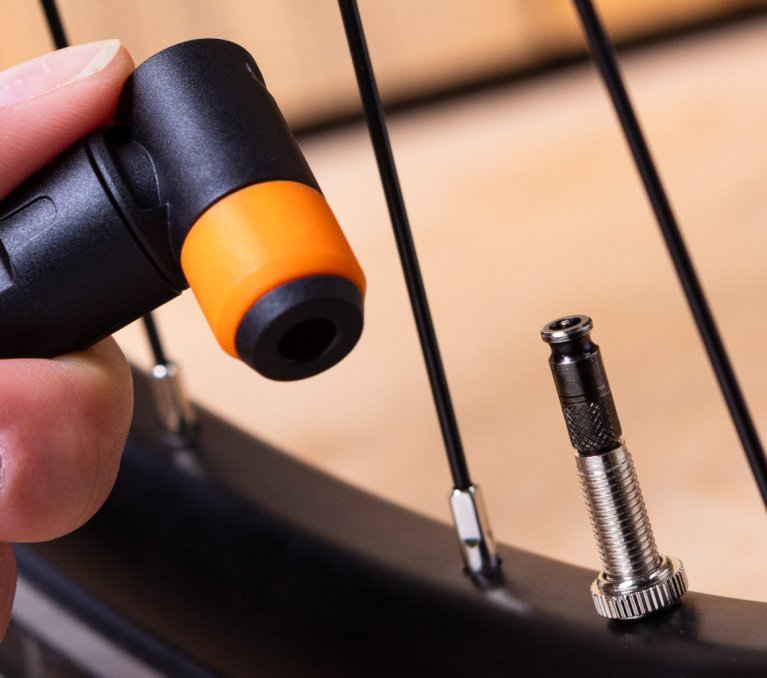 A woman’s hand holds a black and orange pump head next to a Schwalbe Clik Valve bicycle valve on a bike wheel.