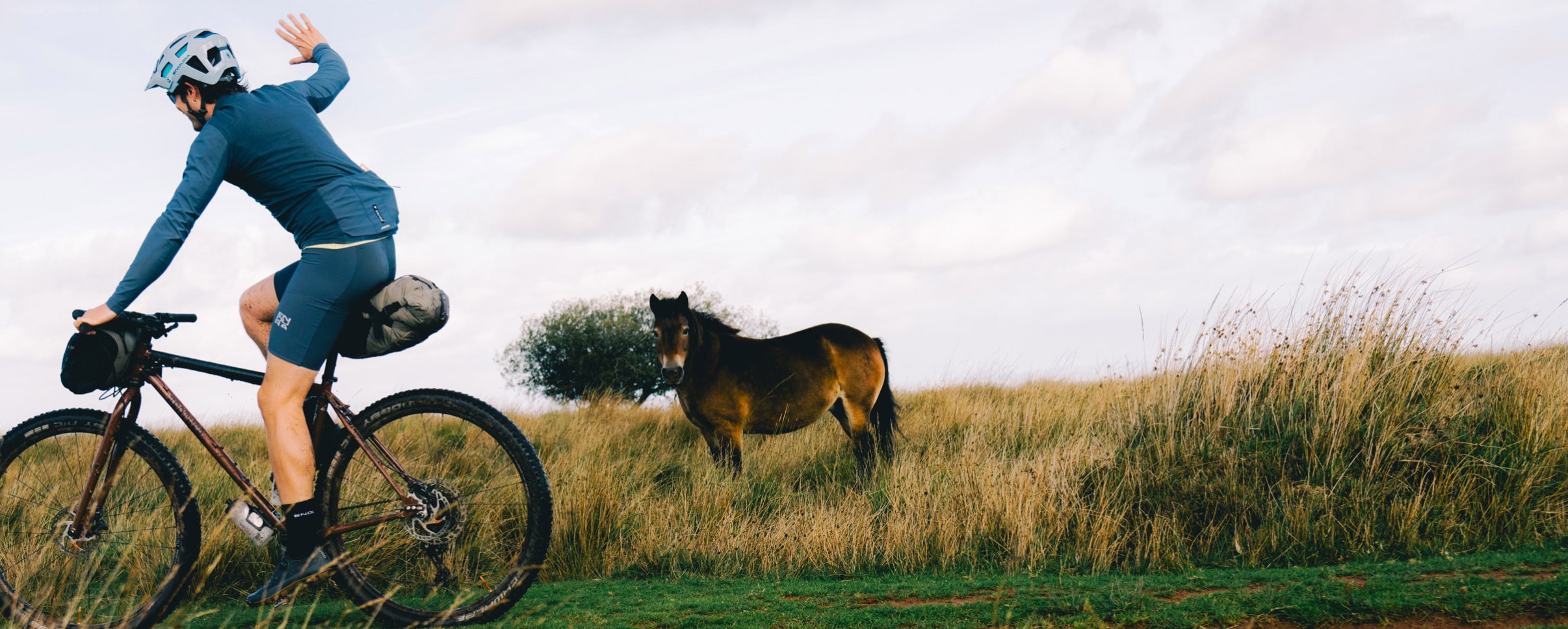 Ciclista de bikepacking Endura en bicicleta gravel saluda a un poni en un paisaje rural de colinas cubiertas de hierba durante una ruta fuera de carretera.