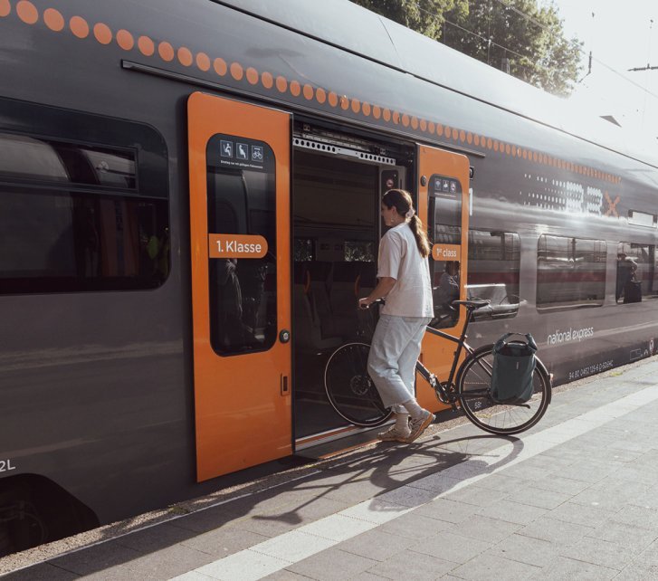 Une personne pousse un vélo équipé d’une sacoche dans un train à quai ; la porte est ouverte et d’autres voyageurs attendent en arrière-plan.