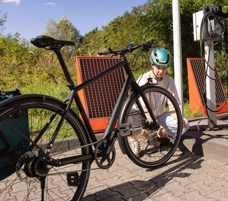 A young woman crouches behind a black city bike and connects the hose of a petrol station compressor to her tyre valve.
