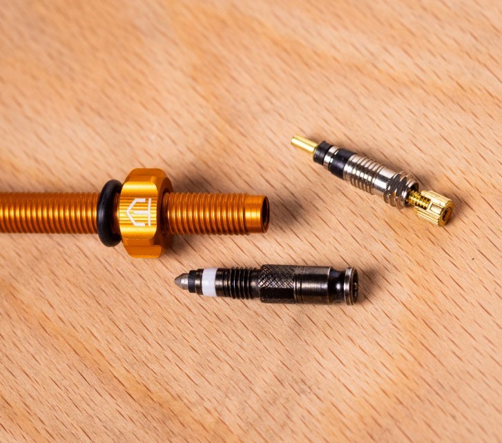 Close-up: An orange anodized tubeless valve lying on a light wooden surface, next to an SV and an SCV valve core.