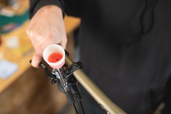 A hand holds a bicycle brake lever with a bleeding funnel attached, filled with red mineral oil, while the background shows a workshop with a wooden table.
