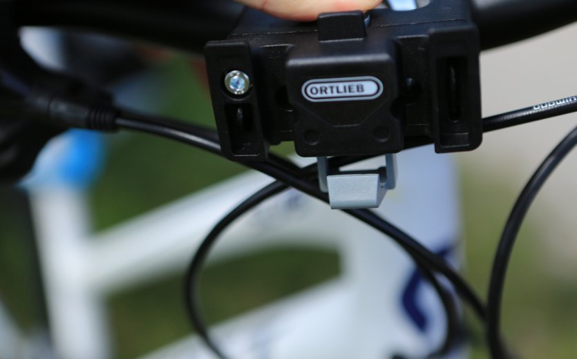 A close-up of the mounting mechanism of an Ortlieb bicycle bag on a bike's handlebar. A blurry green field is visible in the background.