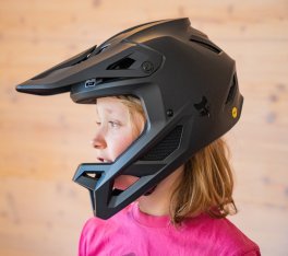 A blonde girl in a pink t-shirt is wearing a full-face helmet for mountain biking.