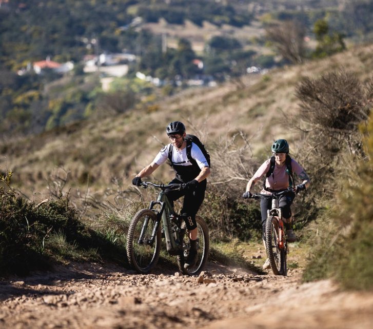 Zwei E-Mountainbiker fahren auf die Kamera zu. Der Trail ist felsig und scheint leicht bergauf zu führen. Sonnenlicht wirft klare Schatten, im Hintergrund ist eine mediterrane, hügelige Landschaft zu erkennen.