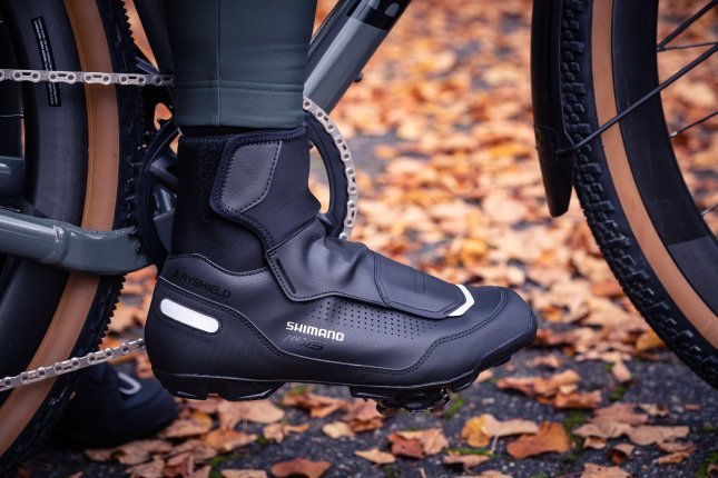 Pie con zapatilla ciclista invernal negra sobre un pedal, hojas otoñales al fondo.