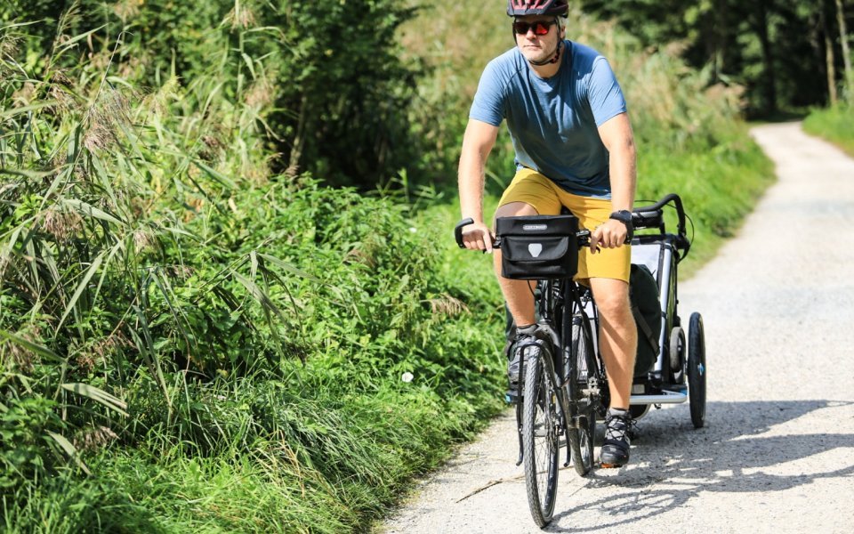 Man cycling with a trailer on a nature path, carrying a black ORTLIEB handlebar bag.
