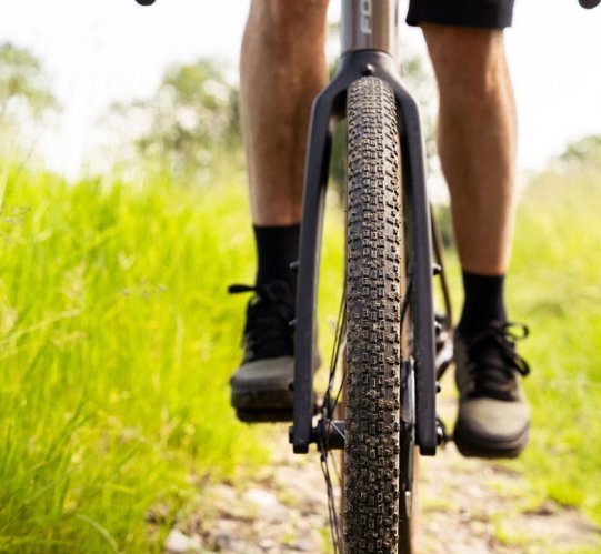 Vue frontale de la roue avant d’un vélo gravel avec pneu à crampons sur un chemin étroit en gravier, entouré d’herbe verte.