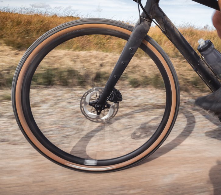 La photo montre la roue avant d'un vélo de gravel bc original Flint en pleine action. Le vélo est en train d'être poussé / roulé sur un chemin de terre. 