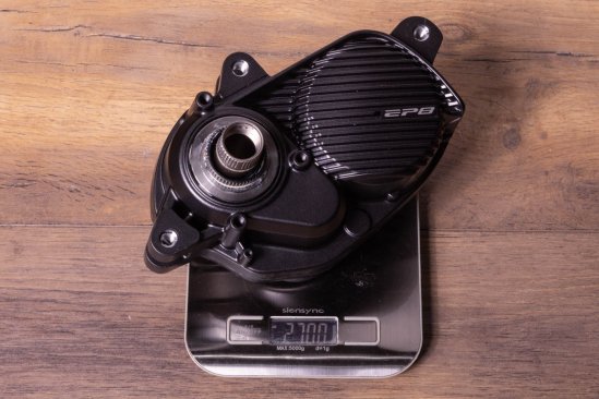 A Shimano EP8 e-bike drive on a kitchen scale on a dark wooden floor. Display shows 2,700 grams