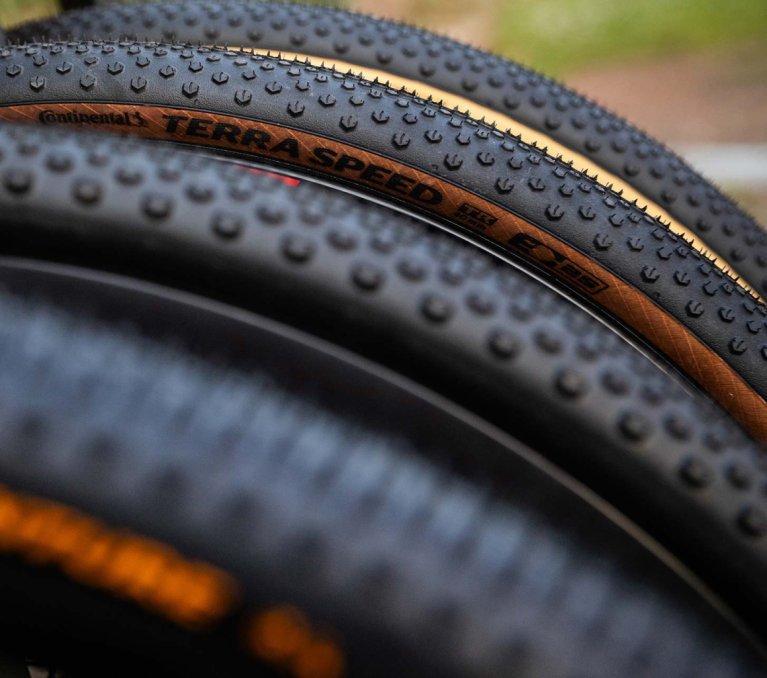 Gros plan de plusieurs pneus de gravel avec profil marqué et flancs beiges, superposés, mettant en avant la structure et la bande de roulement.